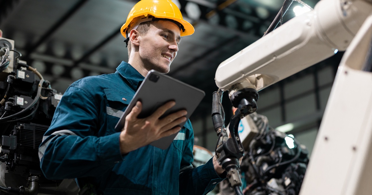 A robotics engineer working on an automated robotic arm inside a warehouse. He is also holding a tablet.