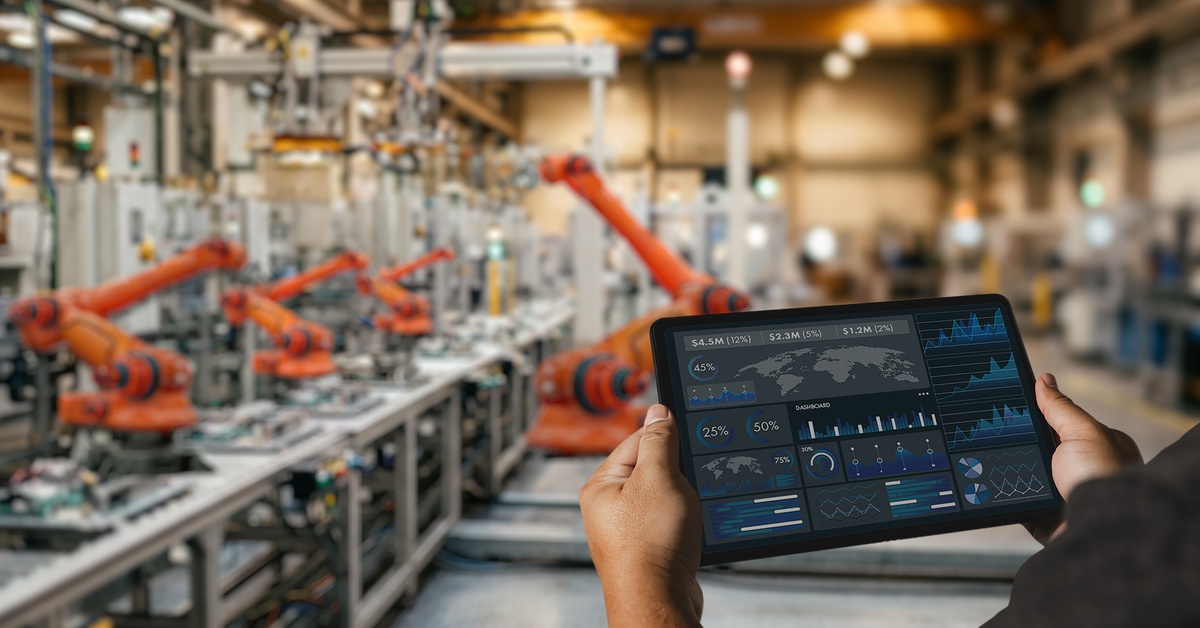 An engineer holding a tablet in a manufacturing factory with robotic automation. The tablet shows analytics and data.