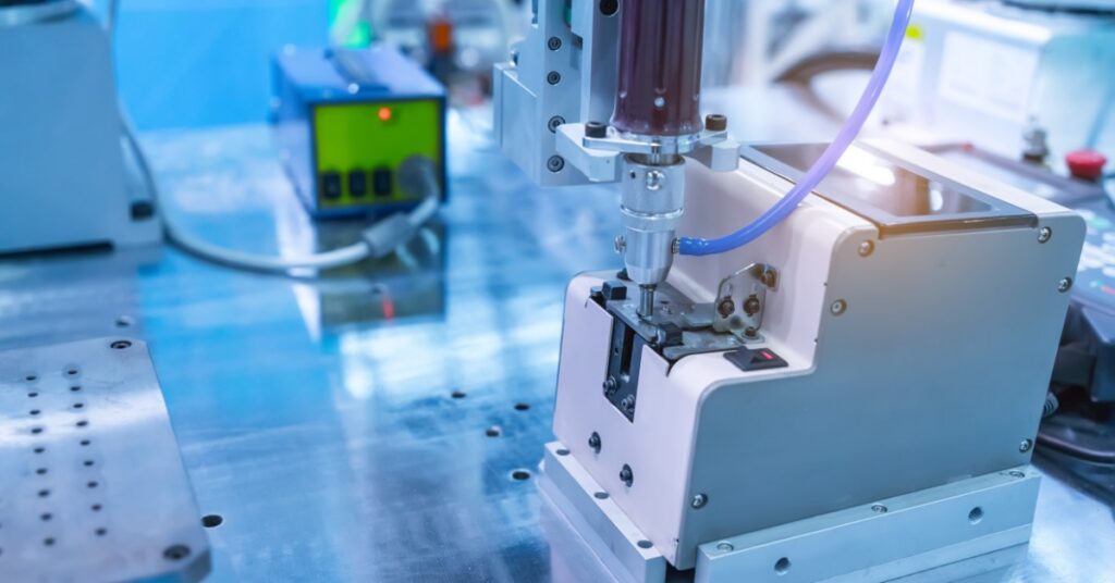 An automatic screw feeder pressing into a small metal fixture on a blue work surface with tubing and equipment nearby.