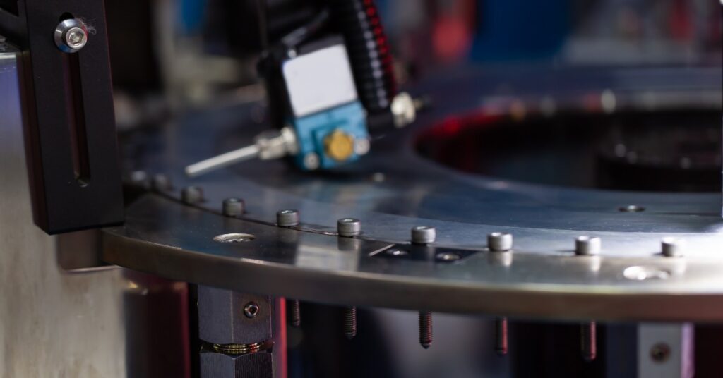 A metal sorting machine with small screws lined along a circular track, sensors, bolts, and dark machinery parts.A metal sorting machine with small screws lined along a circular track, sensors, bolts, and dark machinery parts.