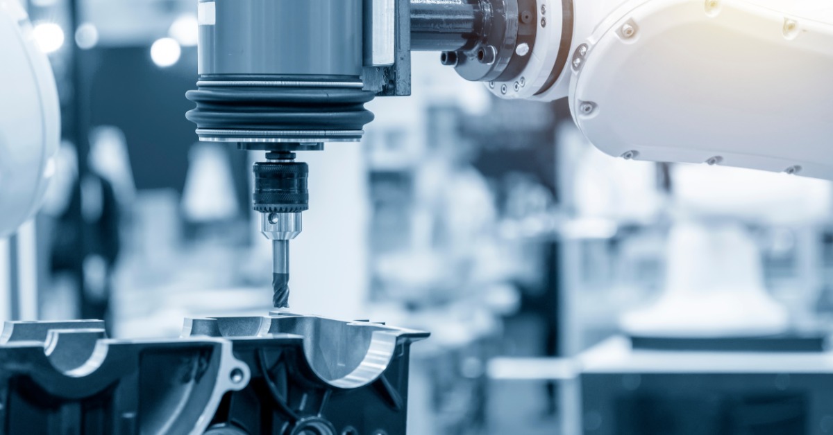 A white robotic arm holds a vertical milling tool over a silver metal automotive part in an industrial workshop.
