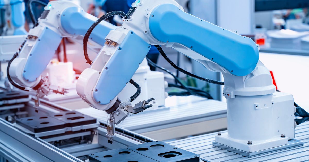 A robotic arm with a small gripper positioned over an electronic assembly line with metal parts and cables nearby.