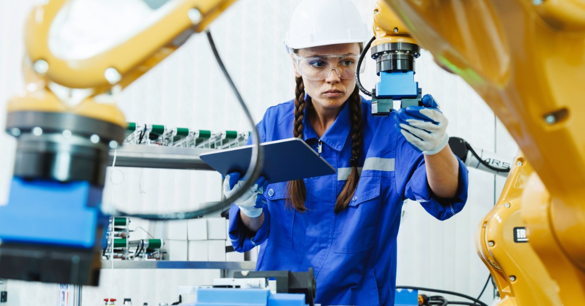 An engineer sitting at a table while using a laptop beside a robotic arm, wires, control equipment, and small parts.